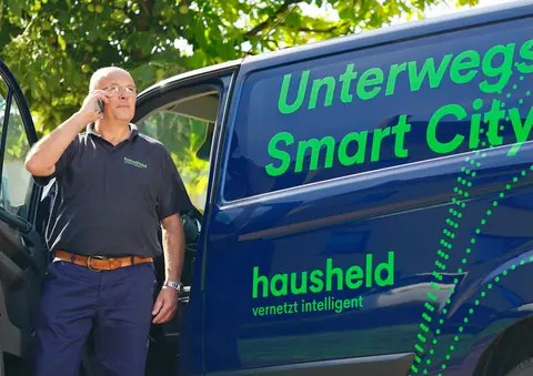 hausheld-Mitarbeiter telefoniert neben einem Firmenfahrzeug mit der Beschriftung „Unterwegs für Smart City“